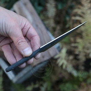 May include: A black and silver metal tool with a sharp, pointed tip. The tool has a dark handle and a shiny, reflective blade. The tool is held against a blurred background of wood and greenery. The tool has the text "SWAYTOBOR TOOLS" on it.