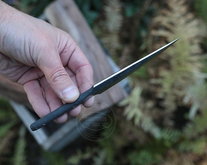 May include: A black and silver metal tool with a sharp, pointed tip. The tool has a dark handle and a shiny, reflective blade. The tool is held against a blurred background of wood and greenery. The tool has the text "SWAYTOBOR TOOLS" on it.