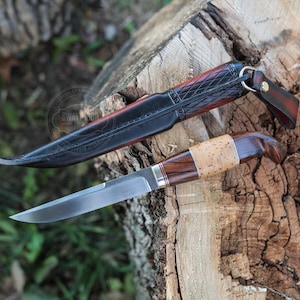Könnte beinhalten: Ein feststehendes Messer mit einer polierten Stahlklinge und einem Griff aus dunklem Holz, einem hellen Birkenrindenabschnitt und roten Zwischenringen. Es wird von einer schwarzen Lederscheide mit Rautenmuster und einem Metallring begleitet.