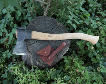 May include: A black and silver axe with a wooden handle, resting on a tree stump. The axe has a leather sheath with the word "SWAYTOBOR" printed on it.