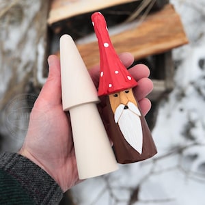 May include: Two wooden mushroom figurines. One is unfinished, the other is painted with a red cap with white polka dots, a brown body, and a white beard. The figurines are held in a hand against a snowy background.