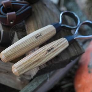 May include: Two wood-handled carving tools with metal loop blades. The handles are light-colored wood with a dark band near the blade. The tools are lying on a piece of wood. The logo "SWAYTOBOR TOOLS" is visible in the background.