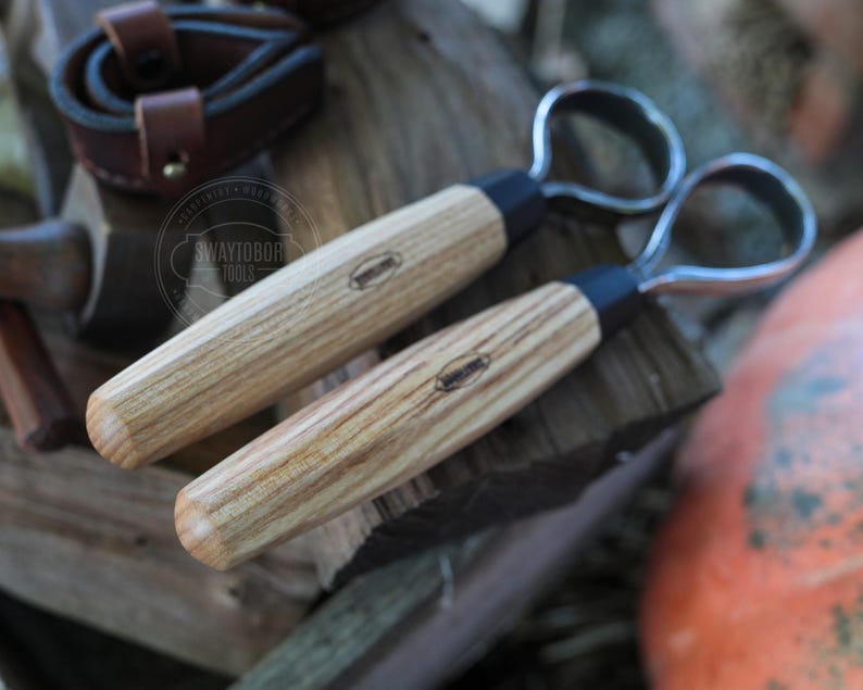May include: Two wood-handled carving tools with metal loop blades. The handles are light-colored wood with a dark band near the blade. The tools are lying on a piece of wood. The logo "SWAYTOBOR TOOLS" is visible in the background.