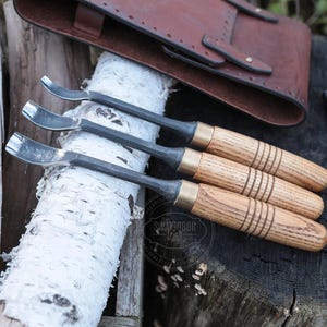 Może przedstawiać: Trzy narzędzia do rzeźbienia łyżek z drewnianymi uchwytami i metalowymi ostrzami są prezentowane na pniu brzozy. Uchwyty mają mosiężne akcenty. Sk&oacute;rzana sakiewka znajduje się w tle.