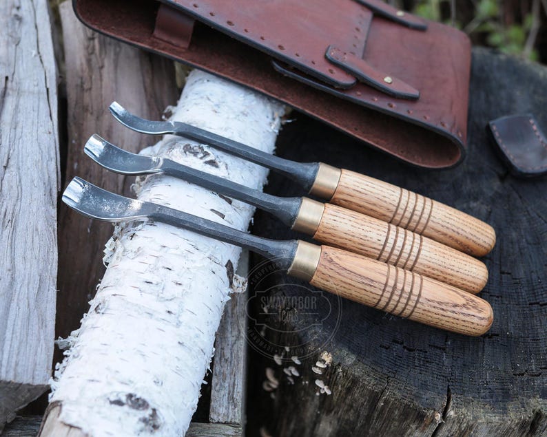 Może przedstawiać: Trzy narzędzia do rzeźbienia łyżek z drewnianymi uchwytami i metalowymi ostrzami są prezentowane na pniu brzozy. Uchwyty mają mosiężne akcenty. Sk&oacute;rzana sakiewka znajduje się w tle.