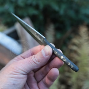 May include: A hand-forged, small, pointed knife with a dark, textured handle. The blade is silver and tapers to a sharp point. The handle has several small, decorative indentations. The knife is held against a blurred green background.