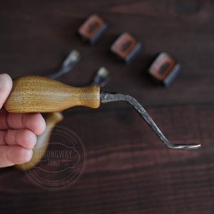 May include: A hand holds a wooden handled, curved, metal tool. The tool is labeled "Strongway Tools" and "Handmade - Forge of Grainers".