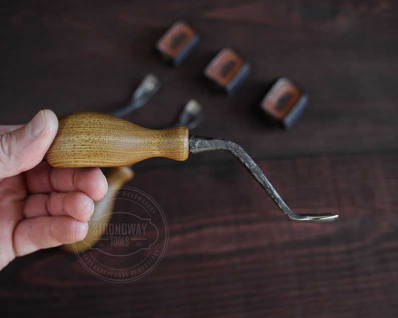 May include: A hand holds a wooden handled, curved, metal tool. The tool is labeled "Strongway Tools" and "Handmade - Forge of Grainers".