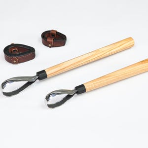Könnte beinhalten: Zwei Hufbearbeitungsmesser mit Holzgriffen und Edelstahlklingen, sowie zwei braune Lederarmbänder mit Schnallen. Die Werkzeuge haben einen schwarzen Kragen, wo die Klinge auf den Griff trifft. Auf weißem Hintergrund.