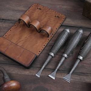 Puede incluir: Un rollo de herramientas de cuero marrón con cuatro ranuras para herramientas. Tres cinceles de mango de madera negro con cuchillas de metal están sobre una superficie de madera.