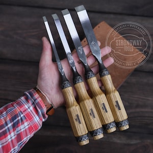 Puede incluir: Conjunto de cuatro cinceles para tallar madera con mangos de madera y hojas de metal. Los cinceles están dispuestos en una mano, con las hojas hacia arriba. Los mangos son todos de color marrón claro y las hojas son todas de color plateado. Los cinceles están etiquetados con "Strongway Tools" y "Handmade - Forge of Britain".