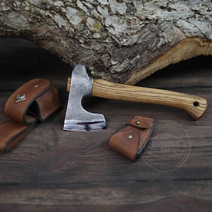 Può includere: Una piccola ascia forgiata a mano con manico in legno e fodero in pelle. L'ascia è appoggiata su una superficie in legno con un pezzo di corteccia dietro.