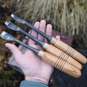 Może przedstawiać: Trzy dłuta do rzeźbienia w drewnie z metalowymi ostrzami i drewnianymi uchwytami. Uchwyty są jasnobrązowe z ciemnobrązowymi pierścieniami. Ostrza są srebrne i zakrzywione. Dłuta są trzymane w dłoni.