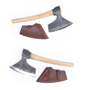 May include: Two hand-forged axes with wooden handles and leather sheaths. The axes have a curved blade and a rounded head. The sheaths are brown leather and have a flap that secures with a snap.