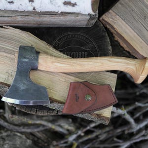 May include: A hand axe with a dark, forged metal head and a light-coloured, curved wooden handle. A brown leather sheath with a metal snap button is positioned below the axe head. The axe features a visible maker's mark on the metal blade.