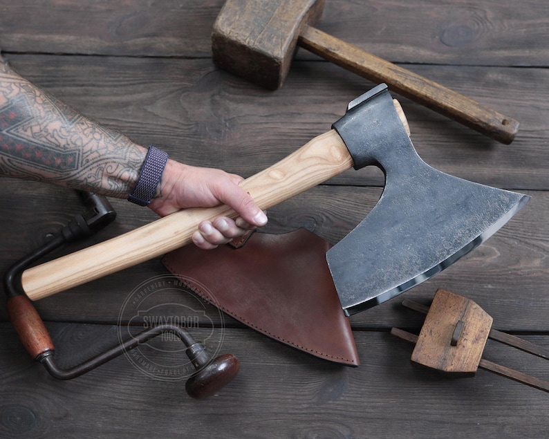 May include: A hand holding a large, curved axe with a wooden handle. The axe has a leather sheath and is resting on a wooden surface. The axe is a traditional style with a wide blade and a curved handle.