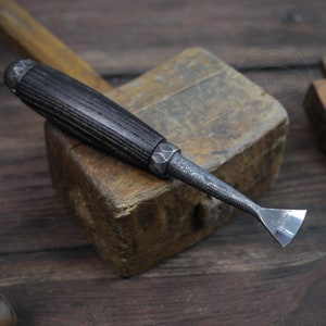 Può includere: Un piccolo strumento per intagliare il legno forgiato a mano con un manico in legno scuro e una lama triangolare affilata. Lo strumento è appoggiato su un mazzuolo di legno.