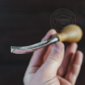 Puede incluir: Un herramienta de talla de madera con una cuchilla curva y un mango de madera. La herramienta se sostiene en una mano y el fondo está desenfocado. La herramienta está etiquetada como "Strongway Tools" con el texto "Carpentry - Woodworking - Handmade - Tools - Experience".