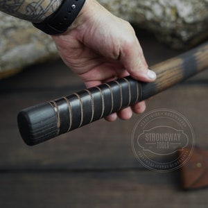 May include: Close-up of a black and brown wooden axe handle with a leather wrap. The handle is held in a hand. The axe head is visible in the background. The axe is labeled "Strongway Tools" with the text "Carpentry - Woodwork - Handmade - Forge of Ukraine".
