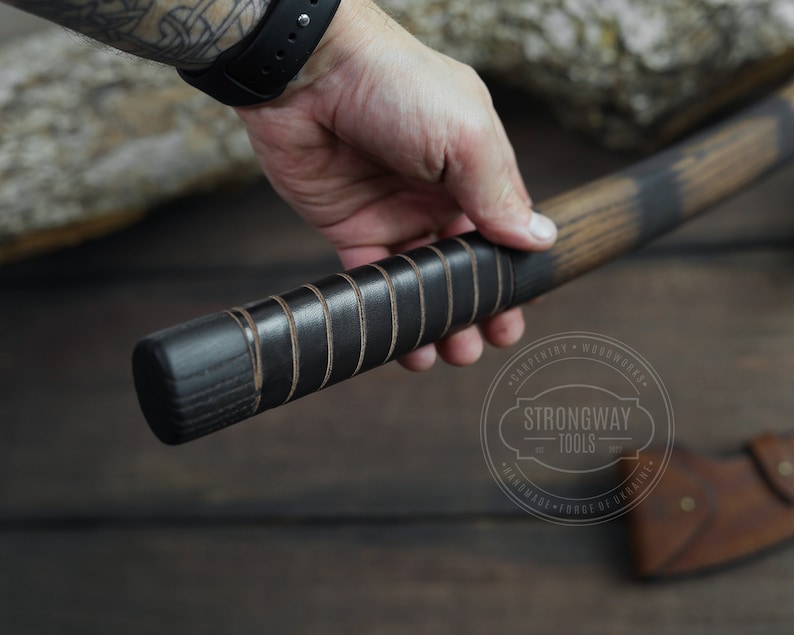 May include: Close-up of a black and brown wooden axe handle with a leather wrap. The handle is held in a hand. The axe head is visible in the background. The axe is labeled "Strongway Tools" with the text "Carpentry - Woodwork - Handmade - Forge of Ukraine".