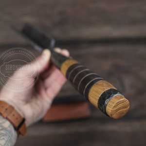 May include: Close-up of a wooden and metal handled tool with a black leather wrap. The handle is made of wood with a dark brown stain. The metal part of the handle is hammered and has a textured surface. The tool is held in a hand with a tattoo. The text "Strongway Tools" is visible in a circle on the background.