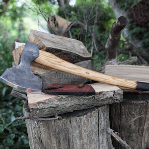 May include: A hand axe with a dark, forged metal head and a light-colored wooden handle featuring a black-dipped end. The axe includes a dark brown leather sheath attached to the blade. The handle has a visible wood grain pattern throughout.