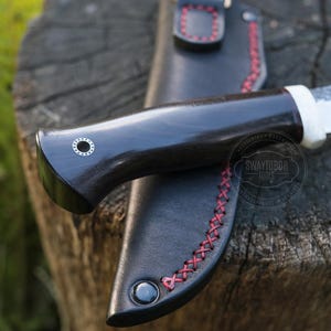 May include: Close-up of a handcrafted knife with a dark wooden handle and a white bolster. The knife rests in a black leather sheath with red stitching. The blade is partially visible, and the knife is set on a wooden surface.