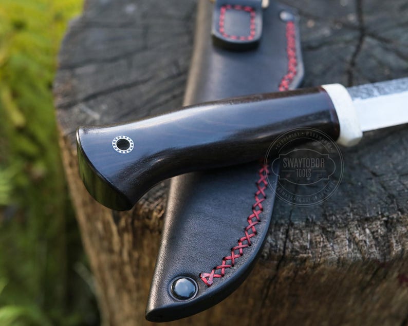 May include: Close-up of a handcrafted knife with a dark wooden handle and a white bolster. The knife rests in a black leather sheath with red stitching. The blade is partially visible, and the knife is set on a wooden surface.
