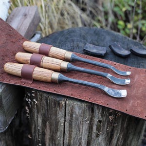 Może przedstawiać: Trzy dłuta do rzeźbienia w drewnie z drewnianymi uchwytami i metalowymi ostrzami spoczywają na brązowym sk&oacute;rzanym pasku. Dłuta są zabezpieczone brązowymi sk&oacute;rzanymi paskami. Narzędzia są wystawione na zwietrzałej drewnianej powierzchni.