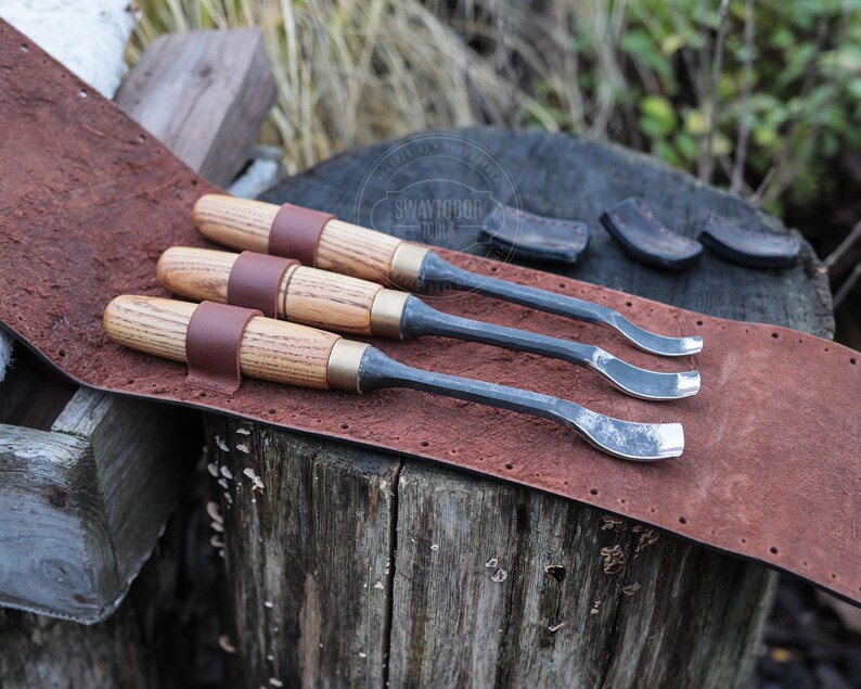 Może przedstawiać: Trzy dłuta do rzeźbienia w drewnie z drewnianymi uchwytami i metalowymi ostrzami spoczywają na brązowym sk&oacute;rzanym pasku. Dłuta są zabezpieczone brązowymi sk&oacute;rzanymi paskami. Narzędzia są wystawione na zwietrzałej drewnianej powierzchni.