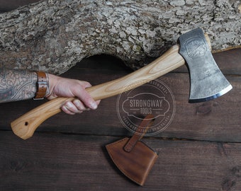 May include: A hand holding a wooden-handled axe with a steel head. The axe has a leather sheath and a logo that says "Strongway Tools" on the handle. The axe is on a wooden background.