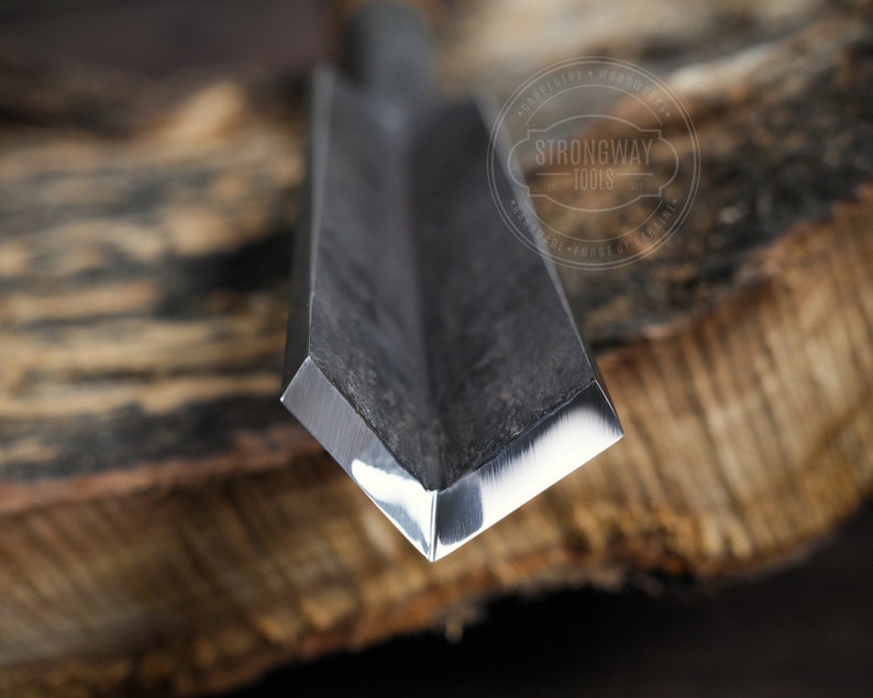 May include: Close-up of the sharp, beveled edge of a metal chisel. The chisel is made of dark metal with a polished, silver-colored edge. The chisel is resting on a piece of wood.