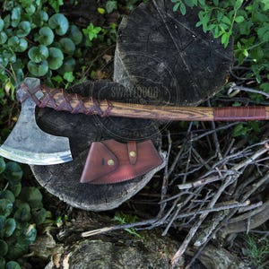 Puede incluir: Un hacha de mano con cabeza de metal oscuro y mango largo de madera envuelto en cordones de cuero marrón. El hacha incluye una funda de cuero marrón a juego con dos botones de presión de latón. La veta de la madera es visible en el mango.