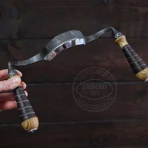 Op de afbeelding: Een gebogen houtbewerkingsgereedschap met twee houten handvatten die zijn omwikkeld met zwart leer. Het gereedschap is gemaakt van metaal en heeft een gepolijst metalen oppervlak. Het gereedschap wordt in een hand gehouden tegen een donker houten achtergrond. De tekst "Strongway Tools" is zichtbaar op een rond label.