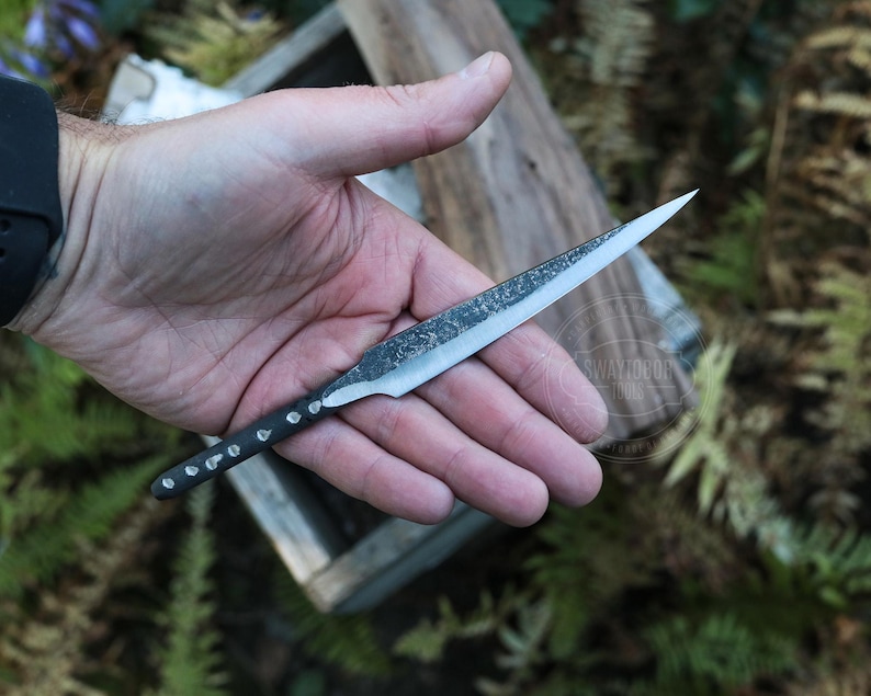 May include: A hand-forged knife with a sharp, pointed blade and a textured, dark metal finish. The handle is black with small, silver-colored rivets. The knife is held against a natural wood background.