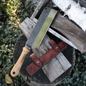 May include: A large cleaver with a wooden handle and a leather sheath. The blade is silver with a gold edge. The brown sheath has a strap with a brass button. The cleaver rests on a wooden surface.