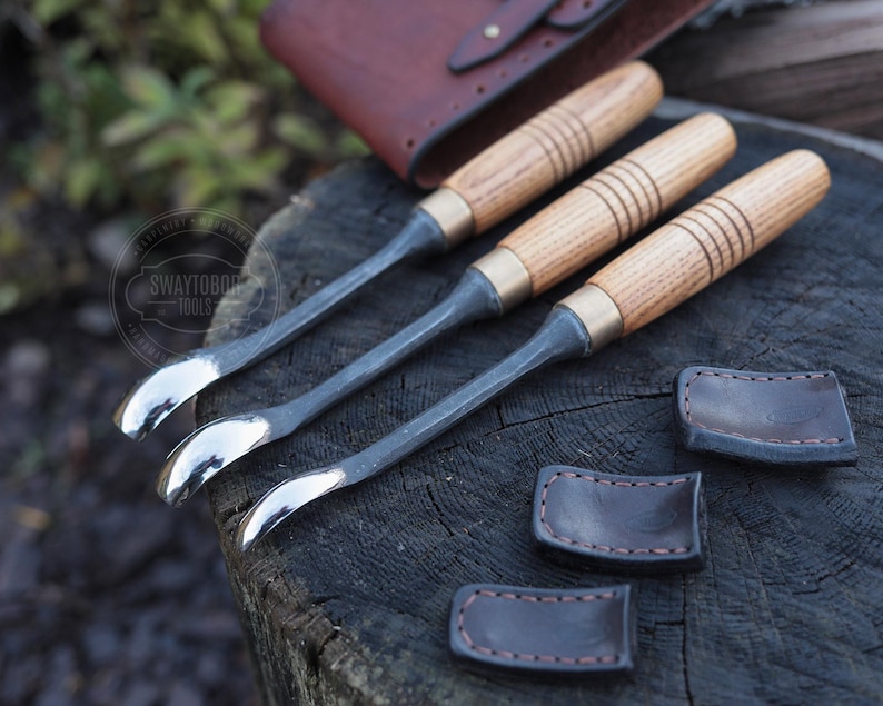 Może przedstawiać: Zestaw trzech dłut do obr&oacute;bki drewna z drewnianymi uchwytami i metalowymi ostrzami. Uchwyty mają mosiężne tuleje i są wykonane z jasnego drewna. W zestawie znajdują się r&oacute;wnież trzy sk&oacute;rzane pochwy, z sk&oacute;rzanym etui w tle.