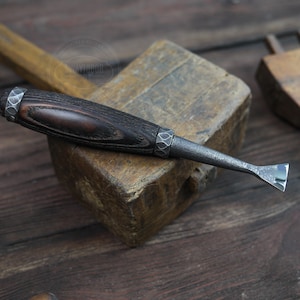 May include: A close-up of a wood carving tool with a dark wood handle and a metal blade. The tool is resting on a wooden block.