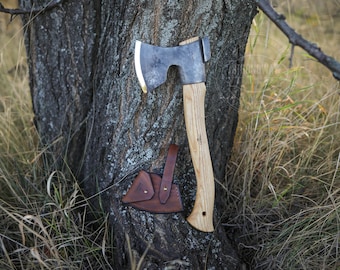 May include: A hand-forged axe with a wooden handle and a leather sheath. The axe is leaning against a tree trunk.