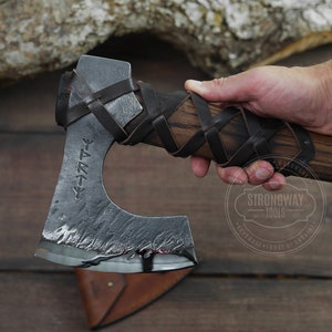 May include: A hand holds a black and brown axe with a leather wrap and a black metal blade. The blade has a runic inscription that reads "Yggdrasil".