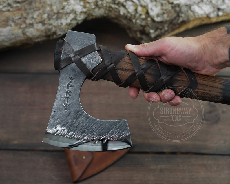 May include: A hand holds a black and brown axe with a leather wrap and a black metal blade. The blade has a runic inscription that reads "Yggdrasil".