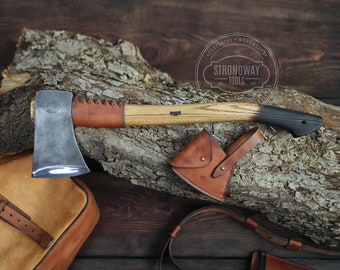 May include: A hand-forged axe with a leather handle wrap and a brown leather sheath. The axe is resting on a piece of wood with the text "Strongway Tools" on a circular label.