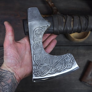May include: A steel axe with a wooden handle wrapped in leather. The axe head features an engraved design. The axe is held in a hand, with the text "STRONGWAY TOOLS" visible. The axe is a tool.