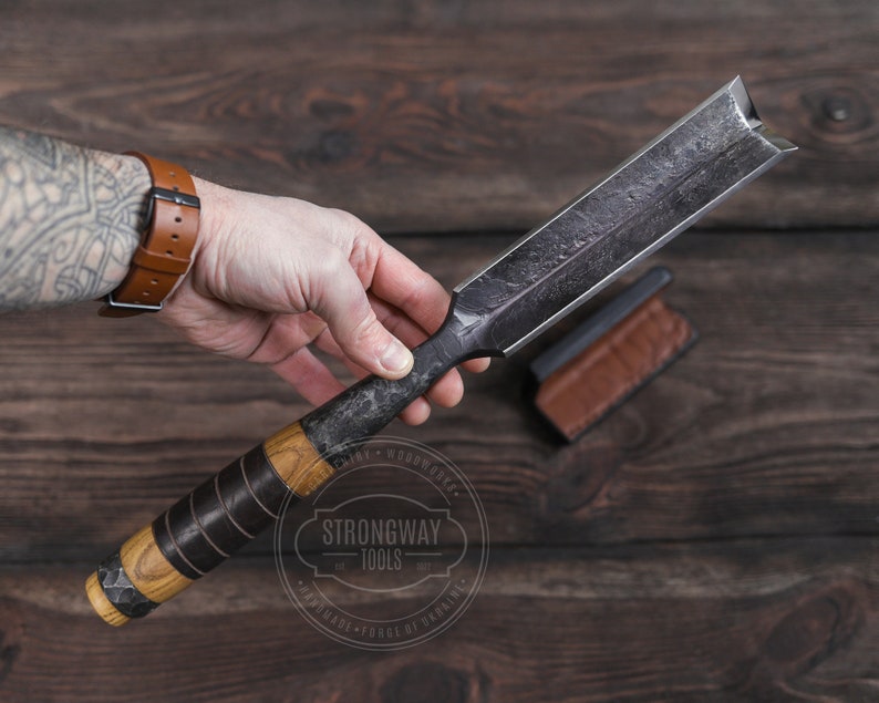 May include: A black and brown wood carving chisel with a leather sheath. The chisel has a wooden handle with a black leather wrap. The chisel is made by Strongway Tools.