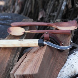 Op de afbeelding: Een houten lepel, een houten spatel en een houtsnijgereedschap met een leren schede liggen op een stapel houtblokken.