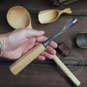 May include: A wood carving tool with a wooden handle and a metal blade. The tool is held in a hand, and the background shows other wooden tools and bowls. The tool is labeled "Strongway Tools" and "Handmade in Ukraine".