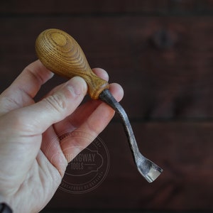 May include: A close-up of a wooden handled carving tool with a curved blade. The tool is held in a hand against a dark wood background. The tool is labeled "Strongway Tools".