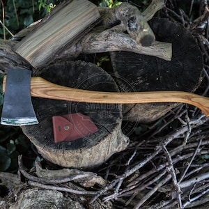 May include: A felling axe with a dark metal head and a long, curved light brown wooden handle. A small, folded brown leather sheath with a metal snap button is positioned directly beneath the axe head on a dark, weathered wooden log surface.
