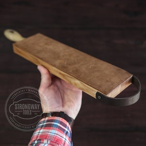 May include: A wooden cutting board with a leather handle. The board is brown and has a natural wood grain. The handle is black and has a metal rivet. The board is being held in a hand.