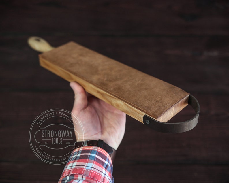 May include: A wooden cutting board with a leather handle. The board is brown and has a natural wood grain. The handle is black and has a metal rivet. The board is being held in a hand.